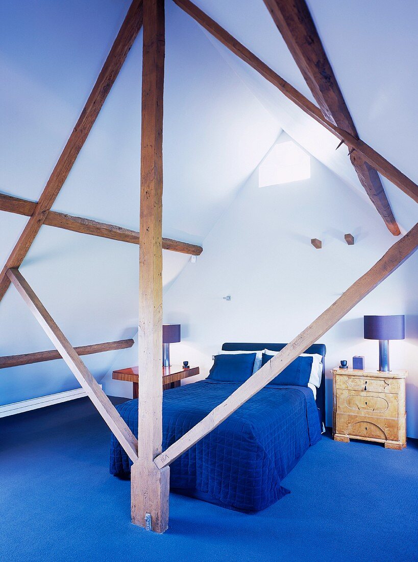 Blick durch Holzkonstruktion auf Bett mit blauer Tagesdecke und blauem Teppichboden unter dem Dach