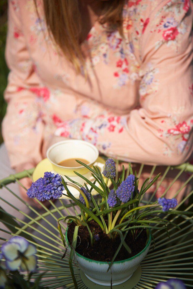 Frau sitzt am Gartentisch mit einer Tasse Kaffee und Traubenhyazinthen