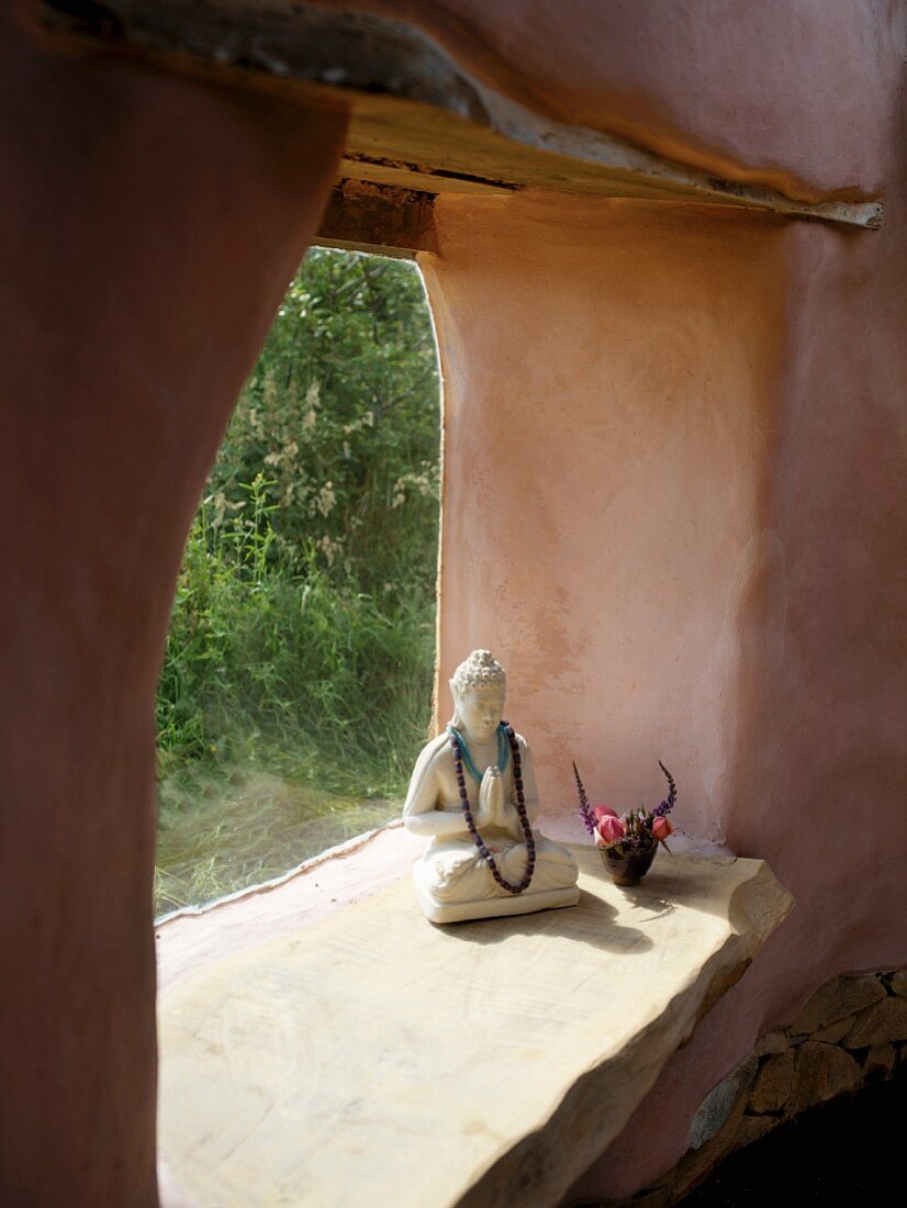 Betender Buddha in der rahmenlosen Fensteröffnung eines organisch geformten Lehmhauses