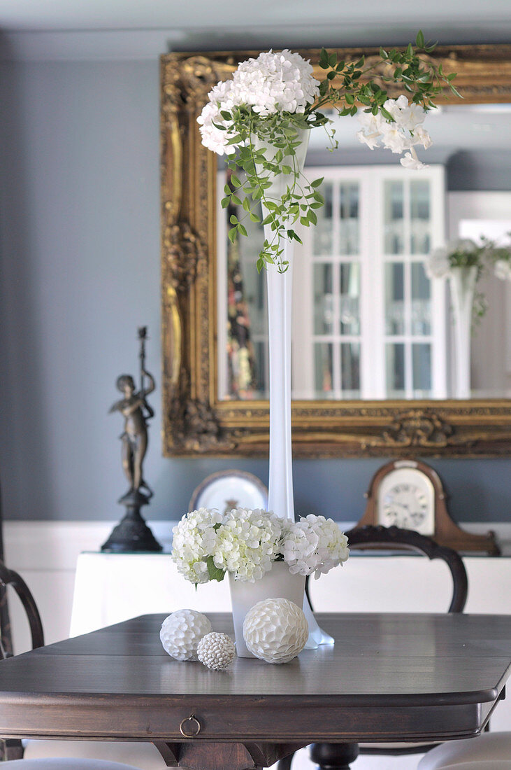 weiße Blumen in verschiedenen Vasen und Dekokugeln auf antikem Tisch vor Spiegel mit Goldrahmen an Wand
