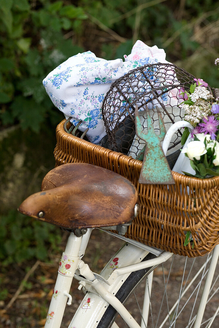An old leather saddle on a vintage white bicycle, with a basket of garden utensils on the pannier rack