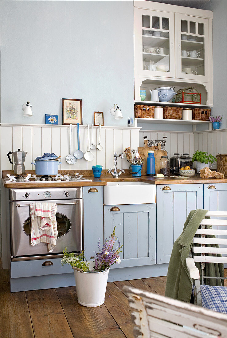 Wooden, country house kitchen counter painted pale blue and white with modern, stainless steel cooker and garden chairs in seating area