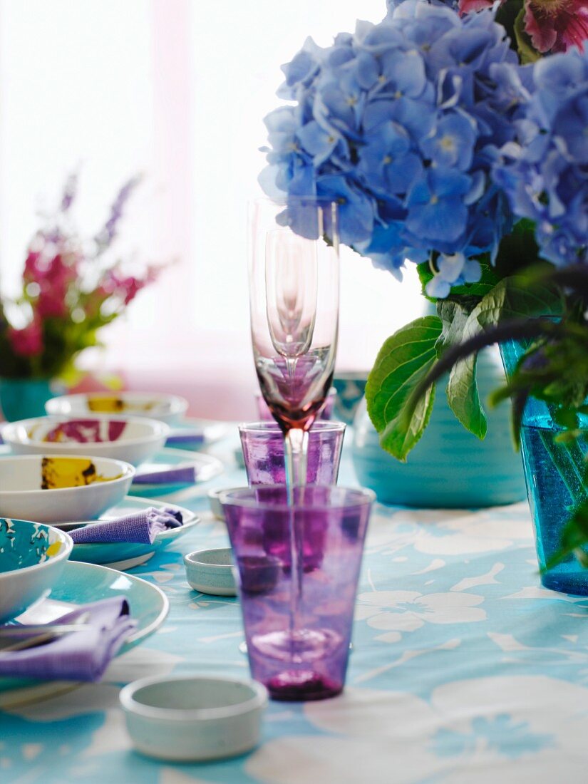 Violette Gläser zwischen Gedecken und Hortensienblumen auf Tisch