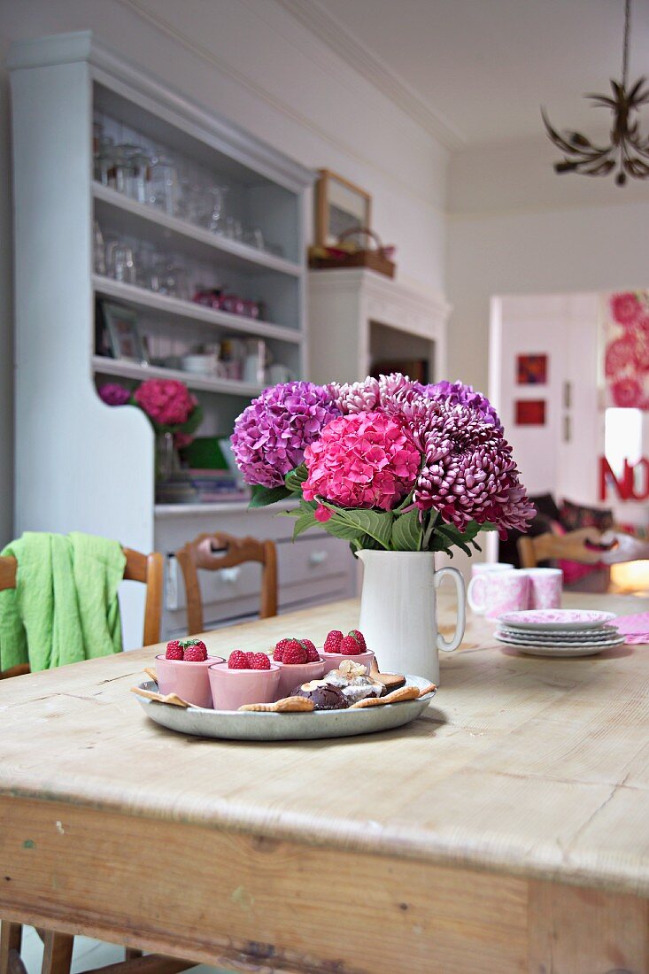 Pink violetter Blumenstrauss im Keramikkrug und Tablett mit Dessert auf rustikalem Esstisch