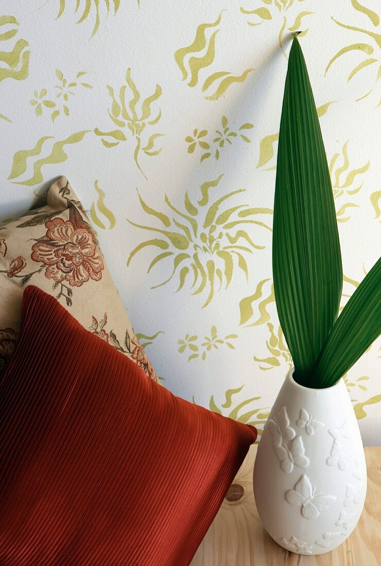 Green leaves in a ceramic vase and decorative pillows in front of floral wallpaper