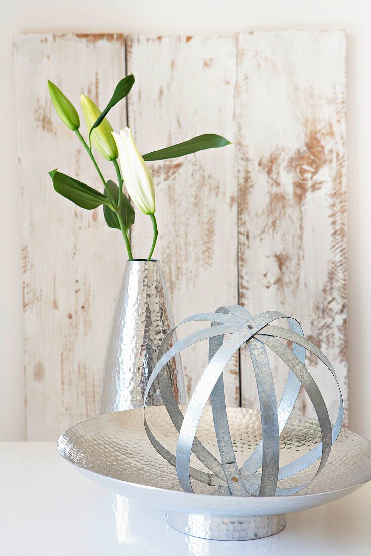 Sphere made of metal strips on metal dish and white lily in metal vase in front of vintage-style wooden board