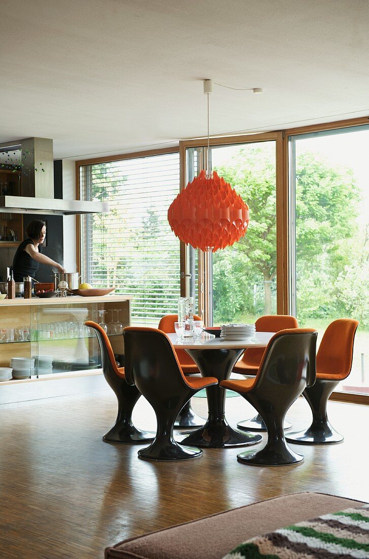 Orangerote Hängelampe über Essplatz mit Schalenstühlen aus braunem Kunststoff und orangen Polstern vor raumhohen Terrassenfenstern