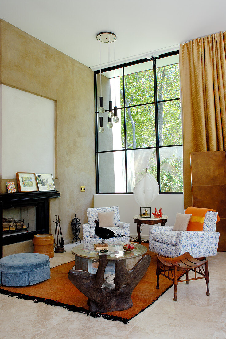 Modernes Wohnzimmer mit Polstersesseln und verschiedenen Hockern vor Kamin und Atelierfenster mit Gartenaussicht