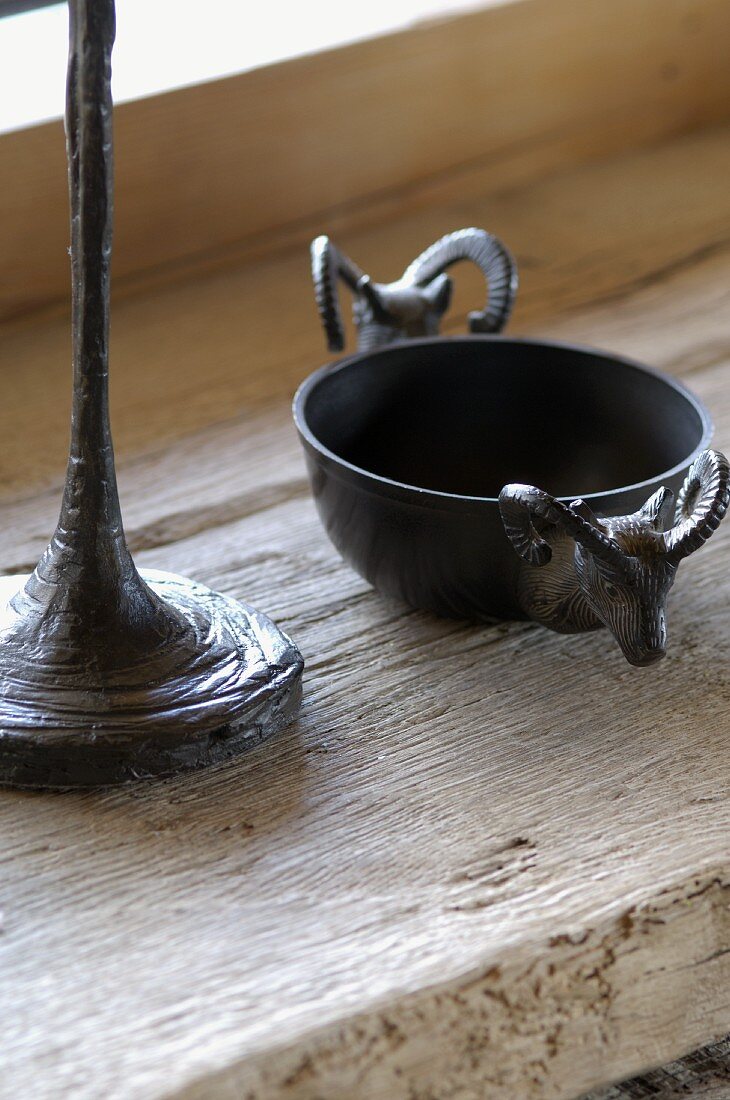 Schälchen aus Metall mit Steinbockköpfen verziert neben Fuss einer Tischlampe auf Vintage Holzablage