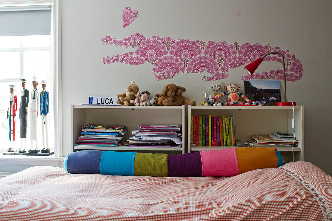 A crocodile and a heart cut out of patterned wall paper above a bed with a colourful relax pillow in a girl's bedroom
