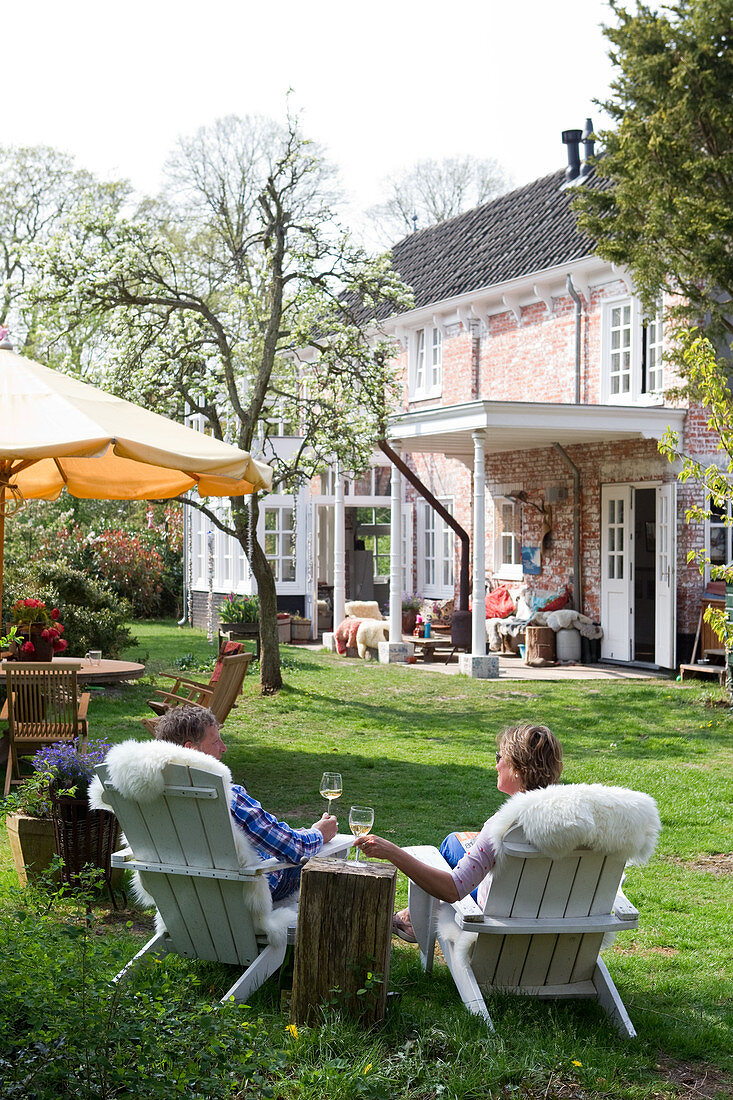 A couple sitting on wooden armchairs covered with sheep skins drinking wine in the garden of a historic Dutch country house
