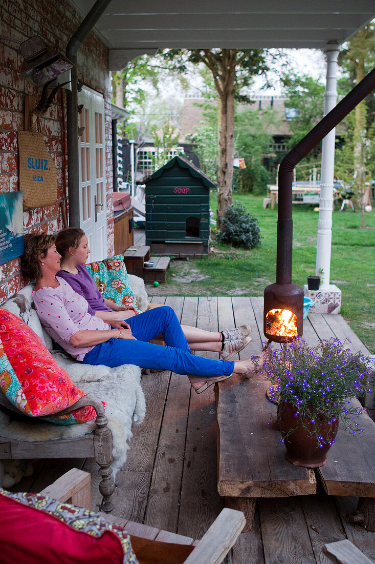 Mutter und Tochter sitzend auf rustikaler Holzbank vor Feuerstelle auf Terrasse
