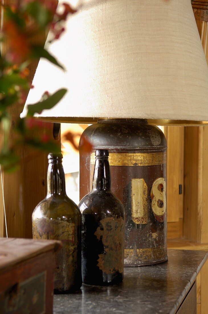 Zwei alte Flaschen als Dekoration neben Tischlampe mit antikem Metallbehälter als Leuchtenfuss