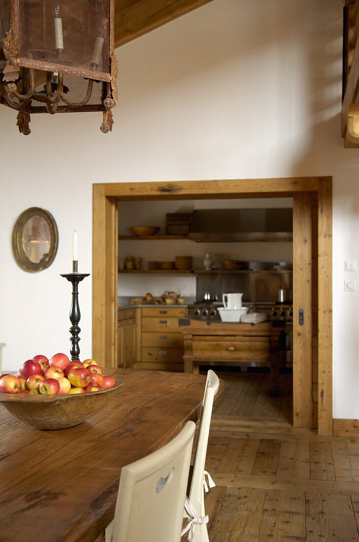 View through a sliding door into the kitchen; a dish full of apples on a rustic dining table and antique lights in a renovated country home