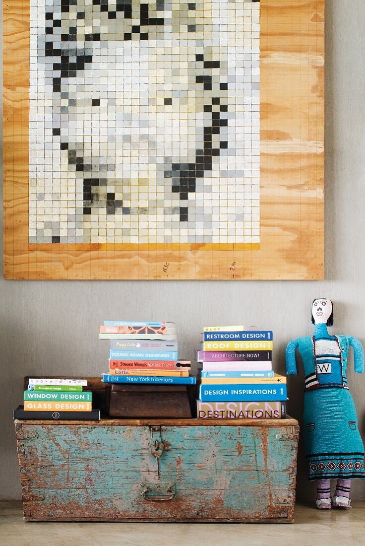 A modern picture hanging on the wall above a stack of books on a vintage wooden chest next to a doll