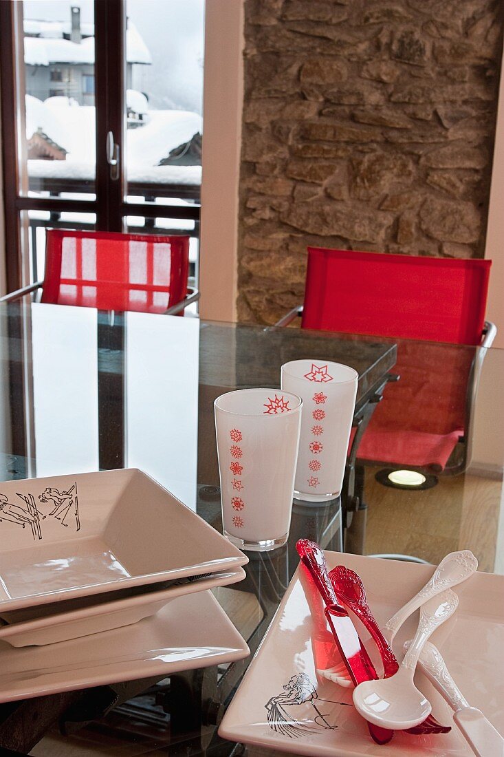 Glastisch mit Essgeschirr und Gläsern vor rustikaler Natursteinwand; im Hintergrund verschneite Berghäuser