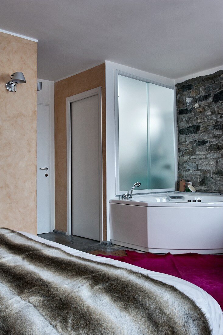 Schlafzimmer mit Eckbadewanne vor rustikaler Natursteinwand; hinter der opaken Glaswand befinden sich Toilette und Waschbecken