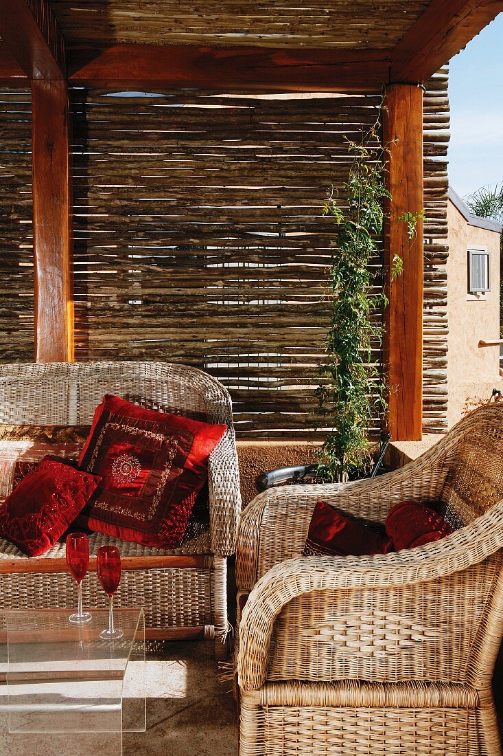 Suite of wicker furniture with red cushions below wooden pergola with bamboo screens