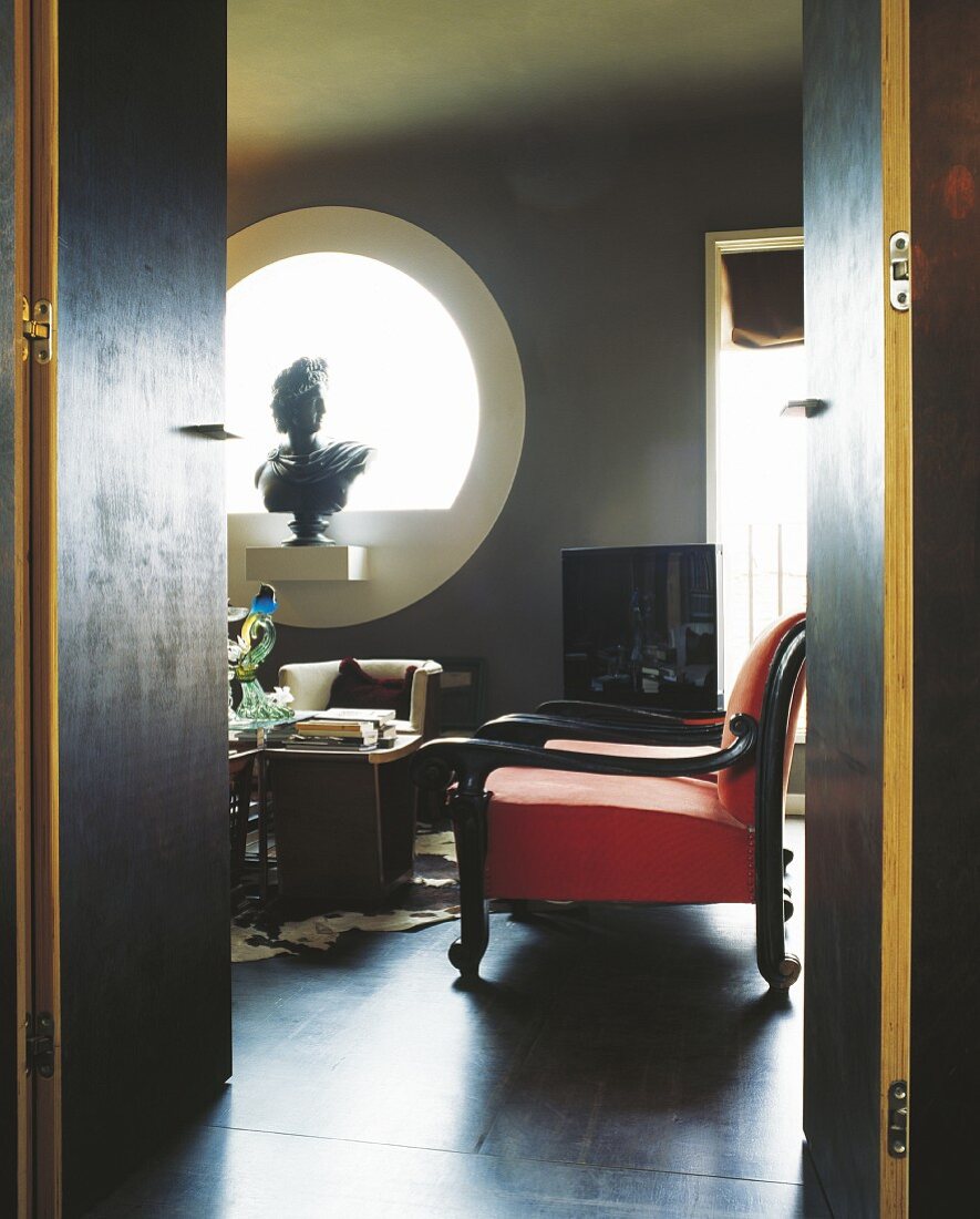 Blick durch offene Tür auf roten Sessel und Skulptur in kreisförmigem Fensterausschnitt in dunkel getöntem Wohnraum