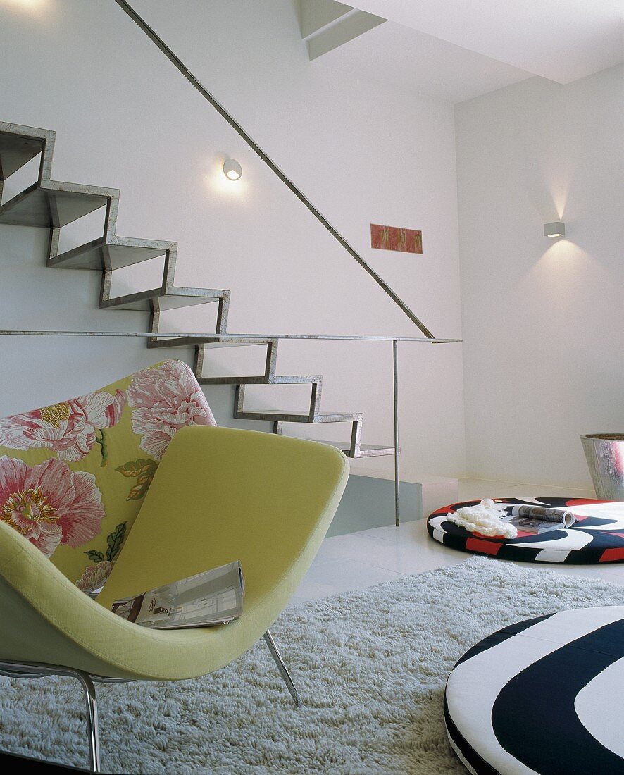 Armchair with green cover in front of modern metal staircase on wall and colourful floor cushions in designer interior