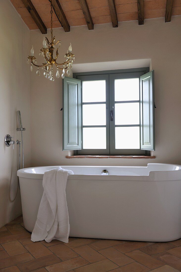 Designer bathtub in front of window and brass chandelier in renovated bathroom of country house