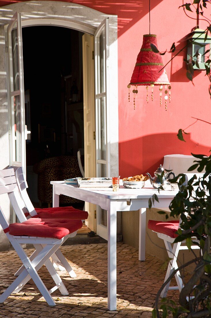Weisser Tisch, weiße Klappstühle mit roten Sitzkissen & rote Hängeleuchte auf teilweise überdachtem Balkon