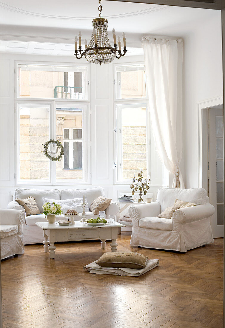 Sofa set with white loose covers and coffee table below chandelier in bay window of living room in period apartment