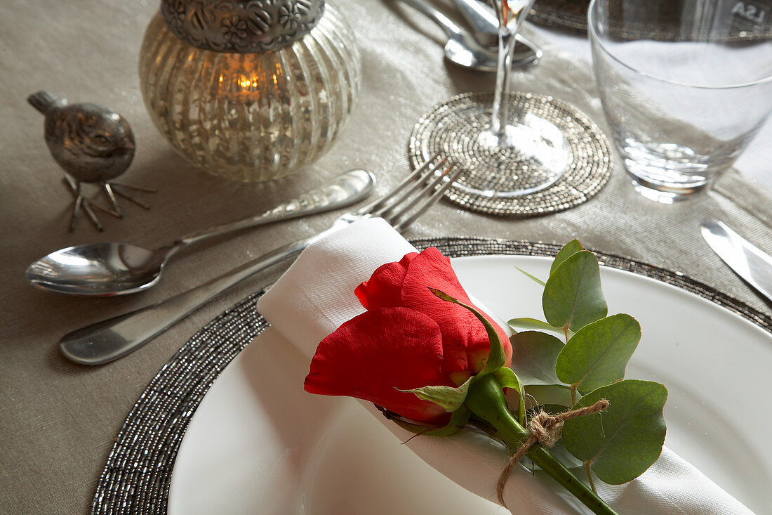 Festliches Tischgedeck mit silberfarbener Deko und roter Rose auf gerollter Serviette