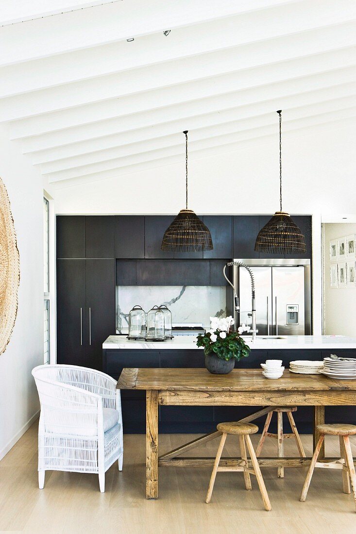 White wicker chair and rustic stools at dining table in front of open-plan, black kitchen