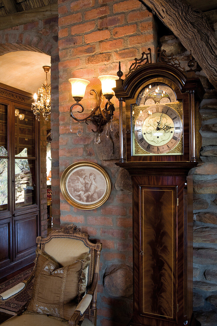 Antique grandfather clock next to upholstered chair and sconce lamp on brick wall