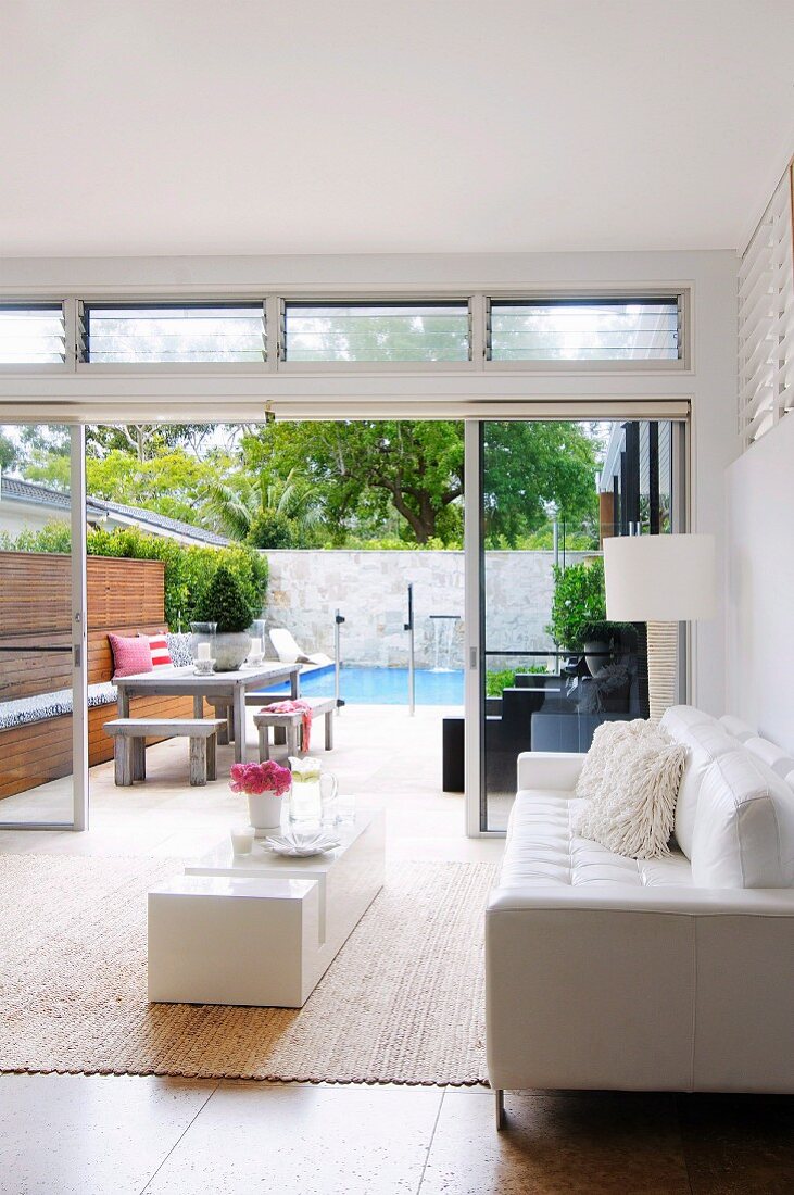 Designer Wohnzimmer mit weißem Ledersofa und minimalistischem Bodentisch vor offener Terrassentür mit Poolblick