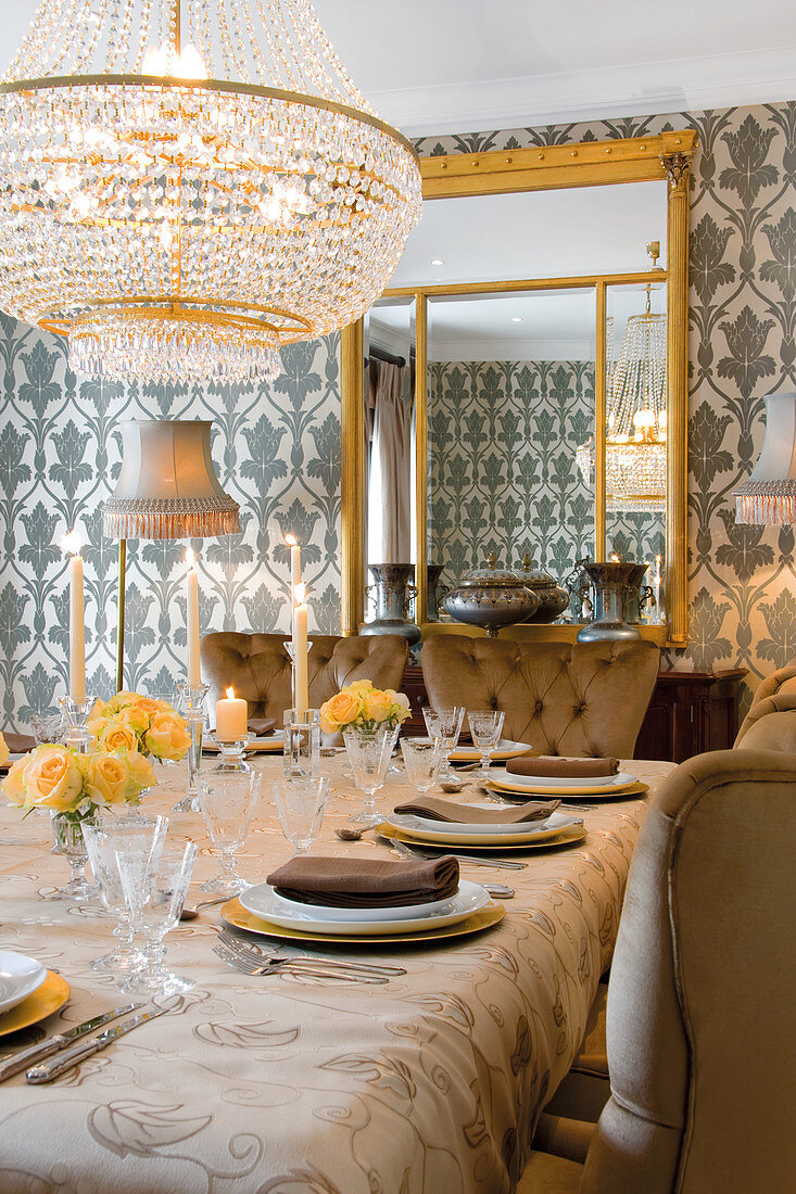 Chandelier above festively set table with tablecloth and upholstered chairs in front of mirror on wall with patterned wallpaper
