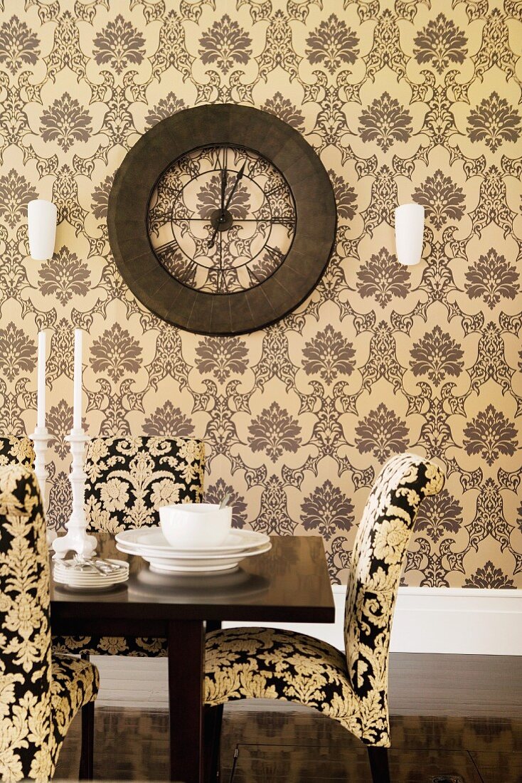 Polsterstühle mit Ornamentmuster am Tisch mit Gedeck vor Wand mit gemusterter Tapete und Vintage Wanduhr