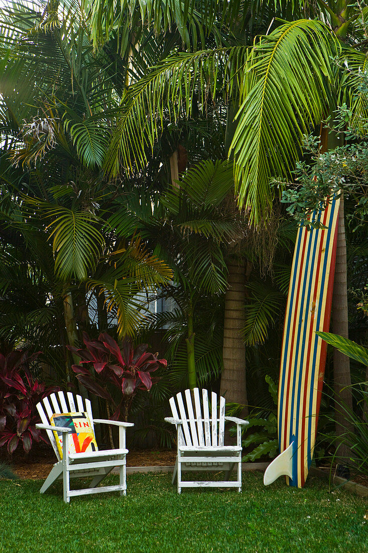 White garden chairs next to a surfboard in the corner of a garden with exotic trees and plants