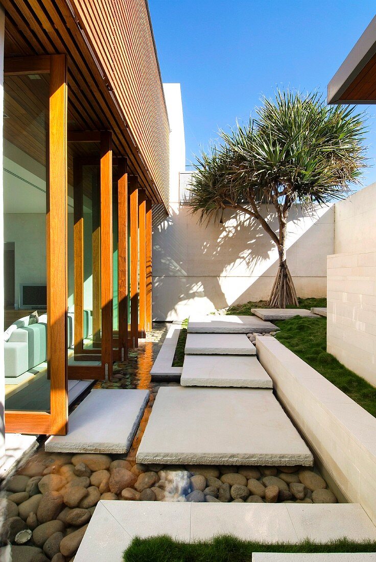 weiße Steinplatten führen über einen flachen Kiesteich vom Wohnzimmer in den gepflegten Garten eines modernen Wohnhauses