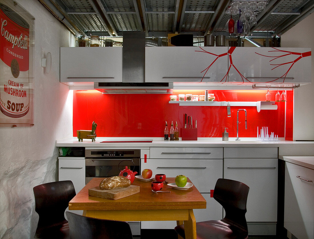 weiße, schlichte Küchenzeile mit leuchtend roter Rückwand und roten stilisierten Ästen auf der Oberschrankfront, Industrie-Decke und gedeckter kleiner Essplatz mit Holztisch im Vordergrund