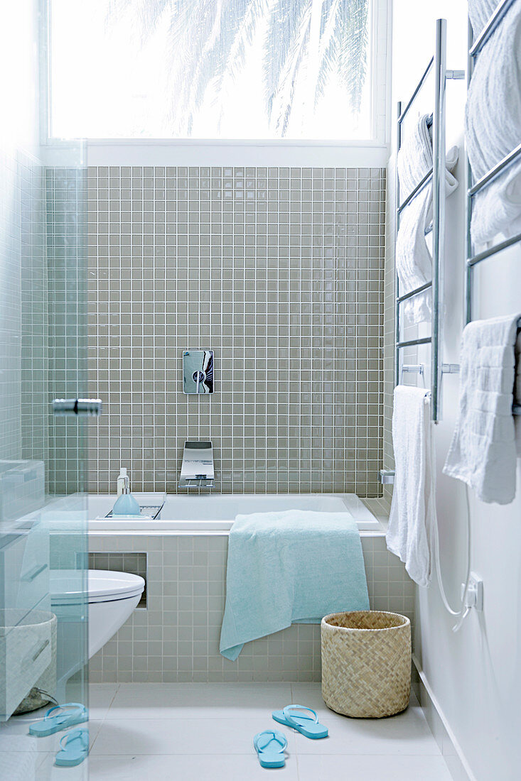View through open glass door of bathroom with bathtub below window, beige wall tiles and stainless steel towel rail