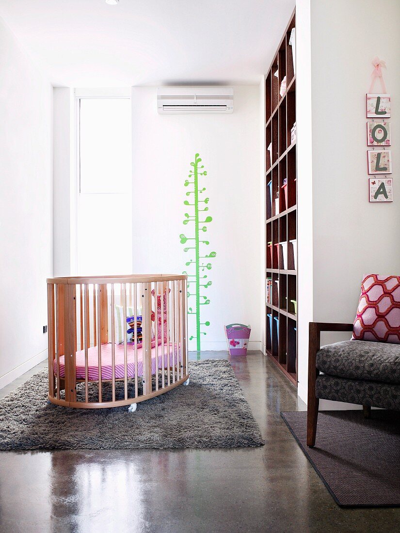 Helles, luftiges Babyzimmer mit Babybett auf weichem Wollteppich und fröhlichem, grünen Baum an der Wand