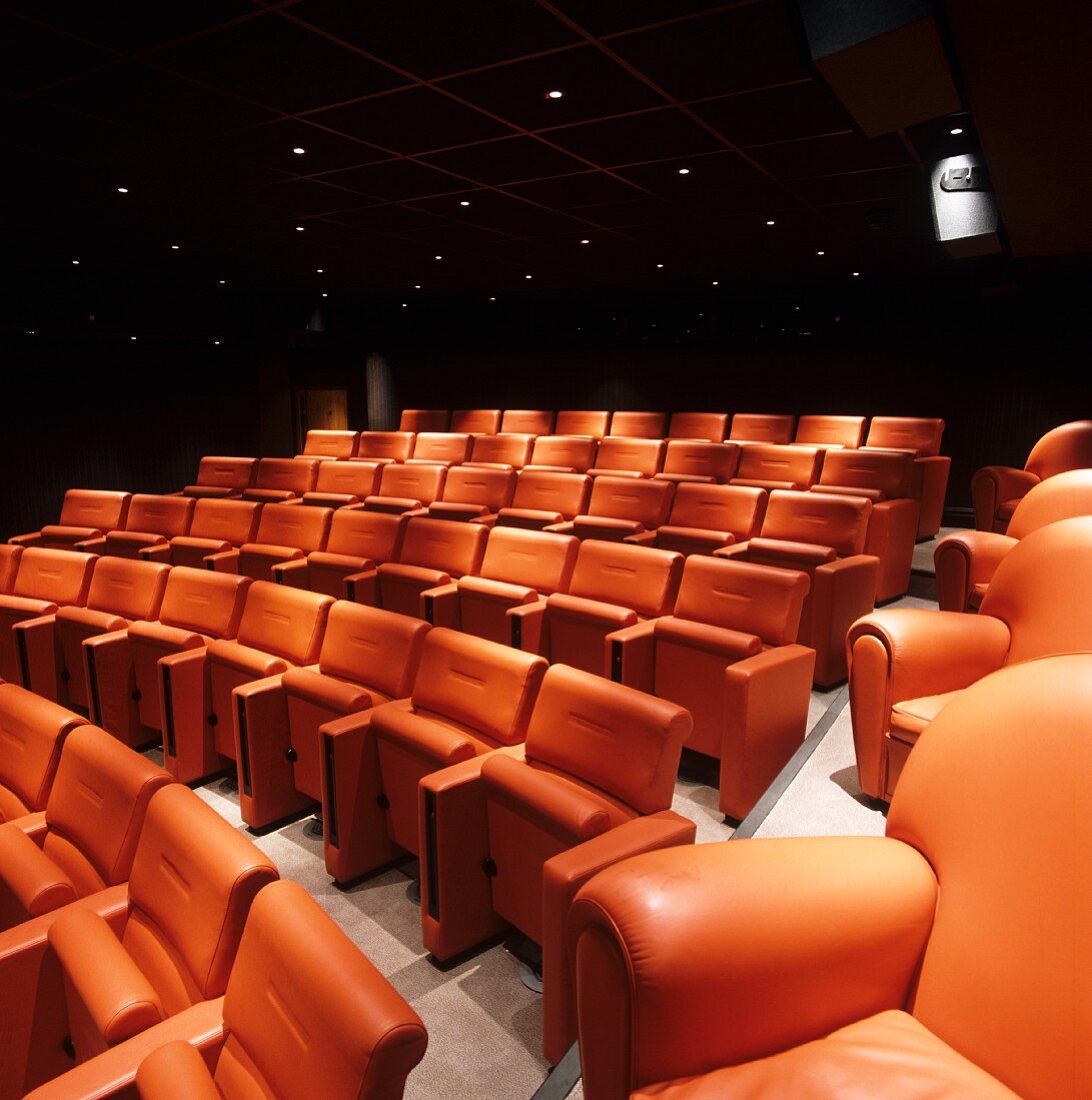 Kinosaal - Stuhlreihen mit orangefarbenen Rücken- und Sitzpolster unter dunkler Kassettendecke mit Strahlern