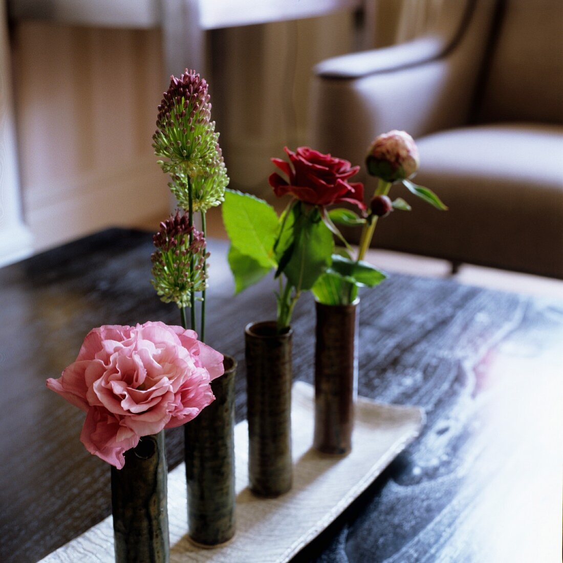 Schmale zylinderförmige Vasen mit einzelnen Blumen auf Schale und Sessel im Hintergrund