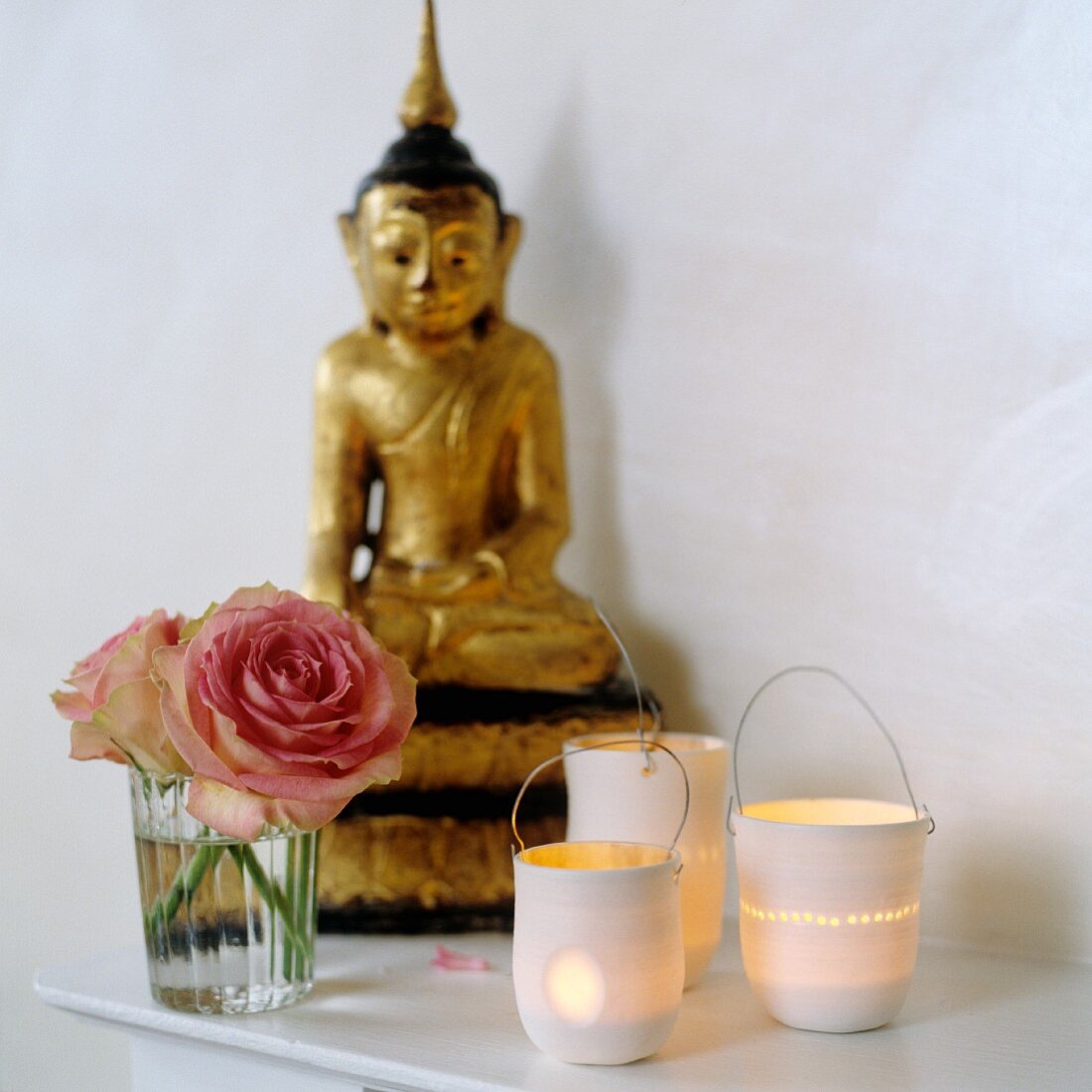 Leuchtende Windlichter neben Rosen im Glas und vergoldete Buddhafigur auf Ablage