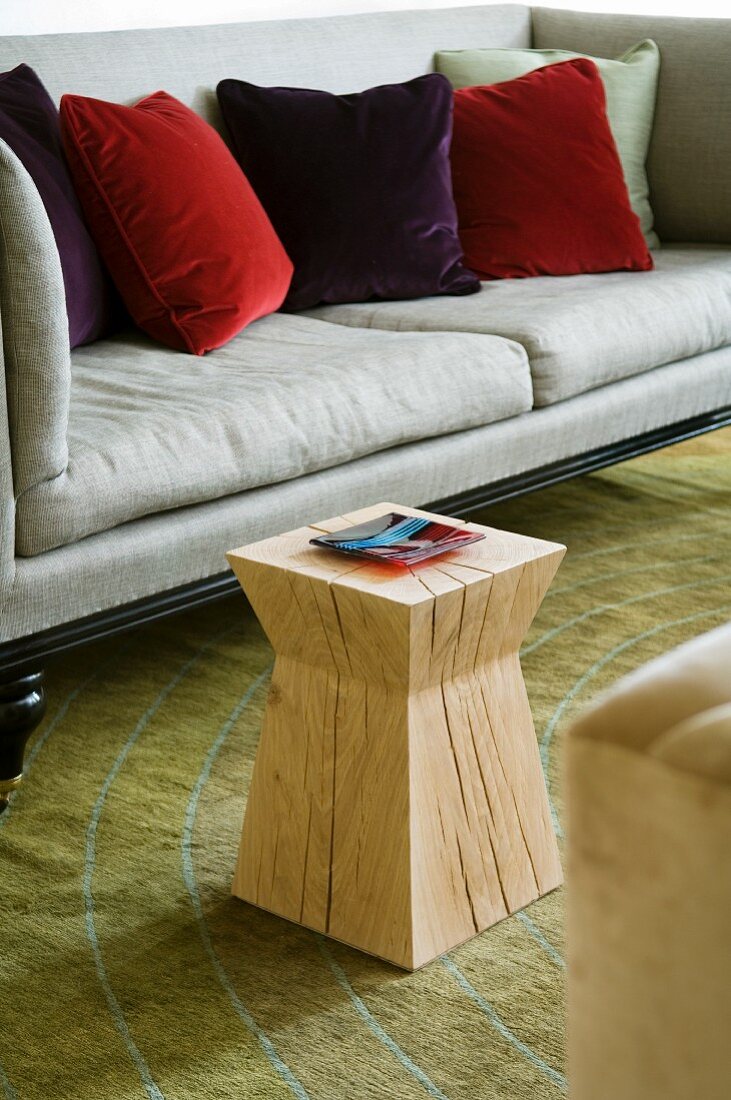 Designerhocker aus Massivholz vor lichtgrauem Sofa mit Samtkissen in Rottönen