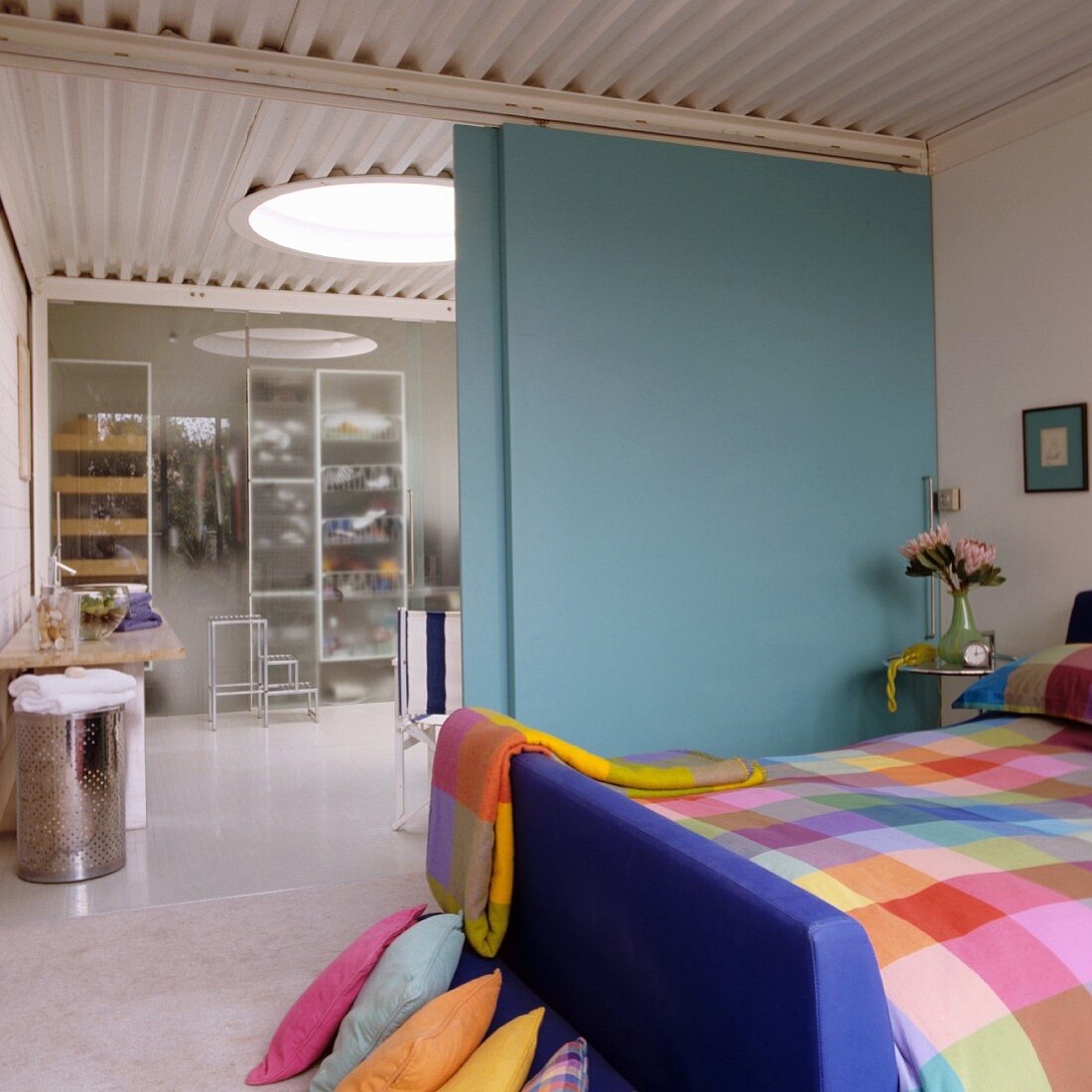 Farbenfroh karierte Bettwäsche im Schlafzimmer und Blick durch eine hellblaue Schiebetür in das Bad Ensuite