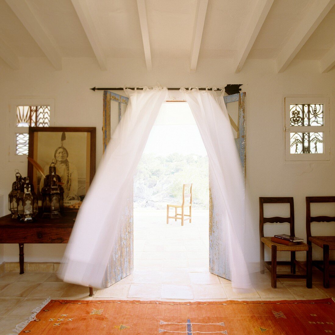 Folkloristischer Teppich vor offener Terrassentür mit luftigem Vorhang und Blick auf Stuhl in Freiem