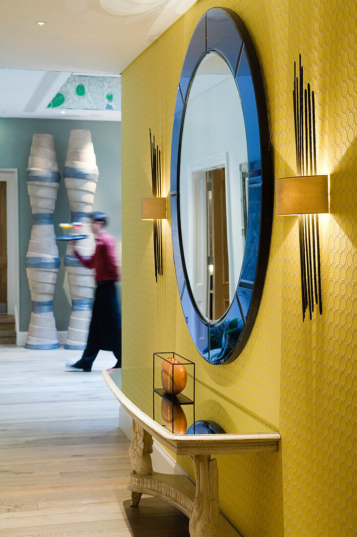 Runder Wandspiegel mit blauem Rahmen zwischen Wandleuchten an gelber beleuchteter Wand in offenem Vorraum und Blick auf Bedienungspersonal