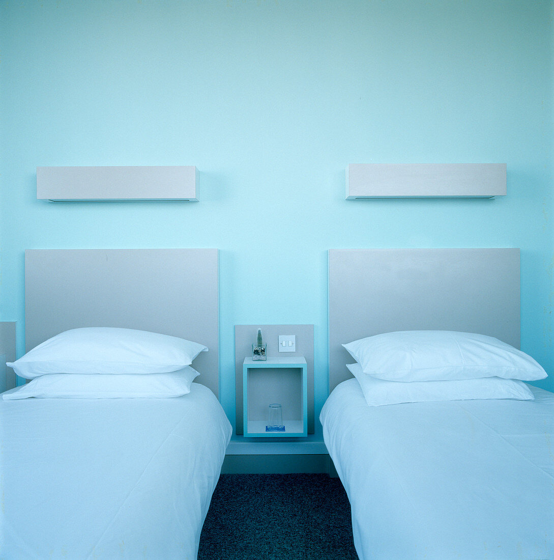 Zwei Einzelbetten mit grau getöntem Kopfteil und Wandbeleuchtung vor blauer Wand in modernem Schlafzimmer