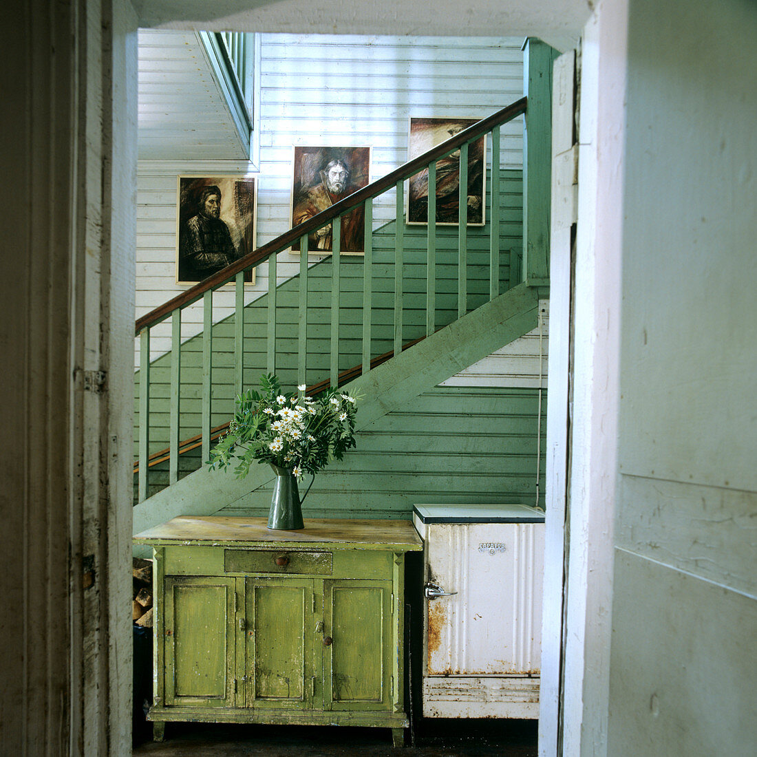 Treppenhaus in grün und weiss mit rustikaler Kommode und rostigem Kühlschrank; frische Blumen sorgen für Fröhlichkeit