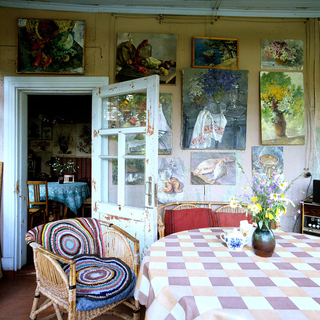 Ölgemäldesammlung an schlichter Holzwand; davor ein Korbstuhl mit runden Häkeldeckchen und ein Tisch mit frischen Wiesenblumen