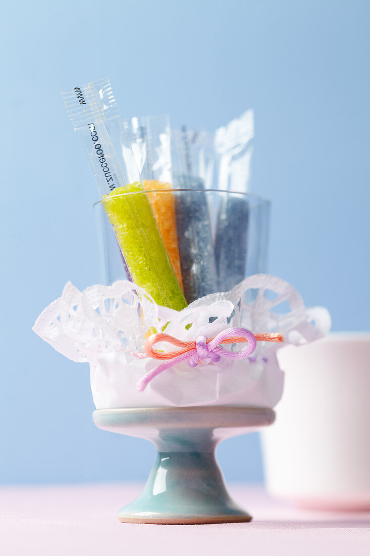 Sticks of coloured sugar in a glass wrapped in a doily on a cake stand