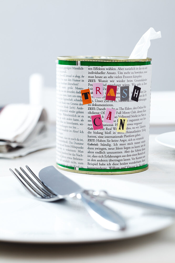 A tin can covered with newspaper and decorated with letters as a table bin